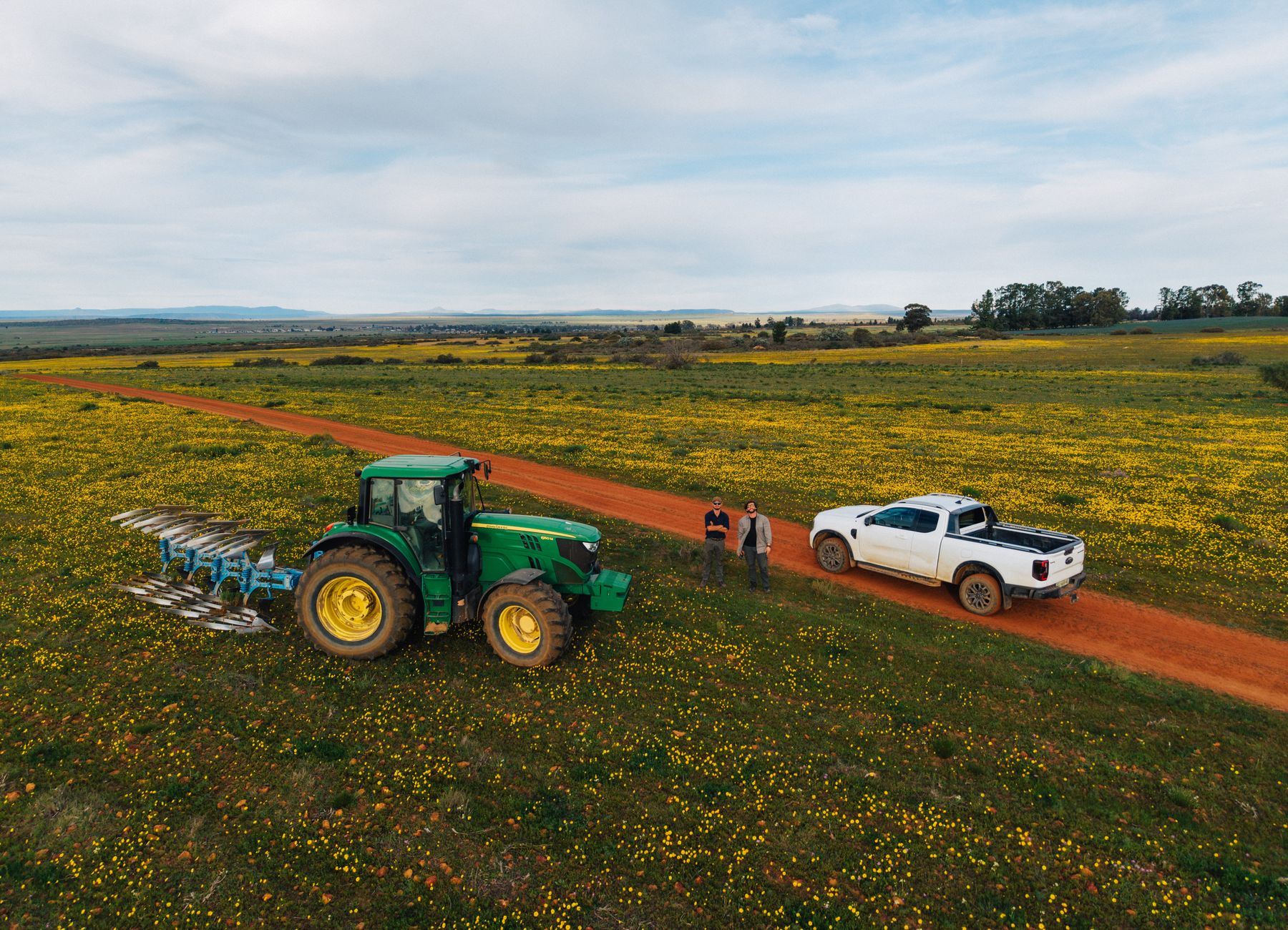 A tractor with an implement and a bakkie on a farm road through yellow spring flowers, from above
